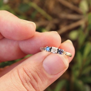 Peut inclure: Une bague en argent ornée d'une pierre centrale transparente, flanquée de deux pierres bleues et de deux petites pierres d'accent. La bague est tenue entre les doigts sur un fond vert flou.
