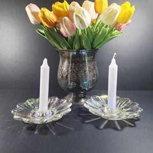 May include: A silver vase filled with colorful tulips, with two clear, iridescent glass candle holders, each holding a white candle. The arrangement is set against a dark background, creating a striking contrast.