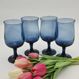 May include: Four blue-tinted glass goblets with dark blue stems and bases are arranged on a white surface. A bouquet of pink and white tulips with green stems and leaves is in the foreground. The goblets are empty.