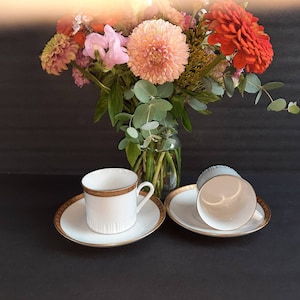 May include: A set of white teacups and saucers with gold detailing. The teacups are on a black surface, with a vase of colourful flowers behind them. The teacups have a classic, elegant design.