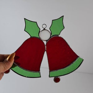 May include: A stained glass Christmas ornament featuring two red bells with green holly leaves and a white bead in the centre.