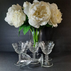 May include: A bouquet of white peonies in a clear glass vase with six clear glass stemmed drinking glasses with etched designs.
