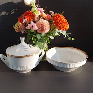 May include: A white porcelain soup tureen with a gold rim and a matching white porcelain bowl with a gold rim. The tureen has a lid and a handle. The bowl is sitting on a black surface. A bouquet of pink, orange, and white flowers is in the background.