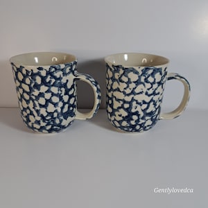 Peut inclure: Deux mugs en céramique avec un intérieur et des anses de couleur crème. L'extérieur présente un motif tacheté bleu et blanc. Les mugs sont présentés sur une surface blanche.