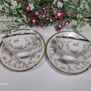 May include: Two white teacups and saucers with pink floral patterns and gold trim. The teacups sit on matching saucers, each with a scalloped edge. The background features white and pink flowers.
