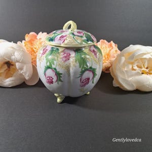 May include: A white porcelain jar with a lid, decorated with pink and green roses and gold accents. The jar has three small feet and is shaped like a pumpkin.