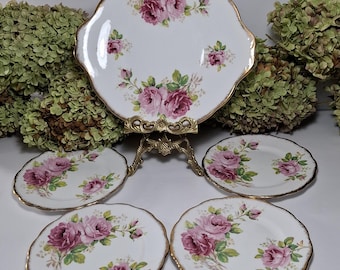 May include: Set of five white porcelain plates with a pink rose floral pattern and gold trim. The plates are arranged on a gold metal stand.