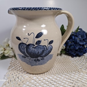 以下が含まれることがあります： 青い縁取りと青と白のリンゴのデザインが施されたクリーム色のセラミックピッチャー。ピッチャーは持ち手があり、白いレースのドイリーの上に置かれています。