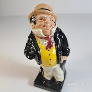 May include: A ceramic figurine of a man wearing a black coat, yellow vest, and a brown hat. He has a white collar and a red tie. The figurine is standing on a black base.