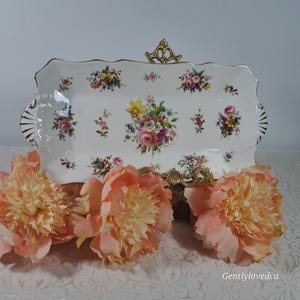 May include: A white rectangular serving tray with a gold rim and a floral pattern. The tray is decorated with a variety of colorful flowers, including roses, daisies, and daffodils. The tray is sitting on a white surface with two peach-colored peonies.