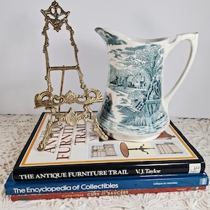 May include: A ceramic pitcher with a blue and white landscape pattern, a small brass easel, and a stack of antique books. The books include titles like "The Antique Furniture Trail" and "The Encyclopedia of Collectibles."