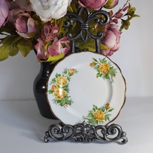 May include: A white porcelain plate with a gold rim and a floral design featuring yellow roses. The plate is on a black metal stand.