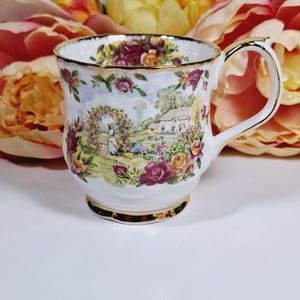 以下が含まれることがあります： 金色の縁と取っ手が付いた白いティーカップ。詳細な花柄と風景のデザインが特徴です。カップは、家、花、人物がいる庭のシーンを描いています。背景にはピンクと黄色の花があります。