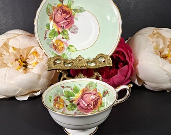May include: A vintage teacup and saucer set with a delicate floral design. The cup and saucer are both white with a green background and feature a pink rose with yellow accents. The set is in excellent condition.