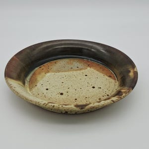 May include: A handmade ceramic plate with a speckled beige centre, transitioning to a rust-coloured band, and a dark brown rim. The plate has a slightly raised edge and a rustic, earthy aesthetic.
