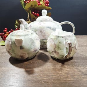 May include: A porcelain tea set comprising a teapot, sugar bowl, and creamer, adorned with a floral pattern in white, green, and purple hues. The teapot features a curved handle and a lid with a white knob. The sugar bowl and creamer have complementary floral designs.