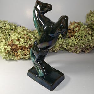 以下が含まれることがあります： 緑色の虹色の釉薬がかかった黒いセラミック製の馬の置物。馬は後ろ脚で立ち上がり、前脚を伸ばしています。置物は長方形の台座の上にあります。