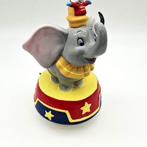 May include: A ceramic Dumbo figurine with a mouse on his hat. The elephant is grey with pink ears and a yellow collar, standing on a red, yellow, and blue circus-themed base with star accents.