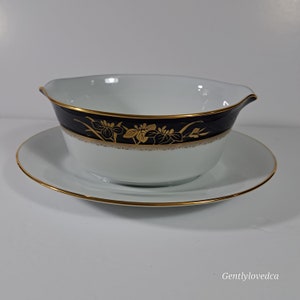 May include: A white porcelain gravy boat with a black and gold rim and floral design. The boat is sitting on a matching saucer.
