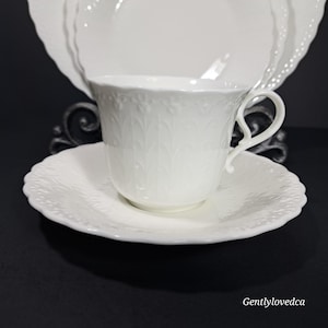K&ouml;nnte beinhalten: Ein wei&szlig;es Tee-Set mit einem zarten Blumenmuster. Die Tasse hat einen Henkel und die Untertasse einen gewellten Rand.