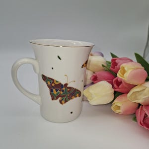 May include: White ceramic mug with a gold rim and a butterfly design in shades of brown, green, and yellow. The mug has a curved handle and is decorated with small green and black dots. A bouquet of tulips in pink, yellow, and white is next to the mug.