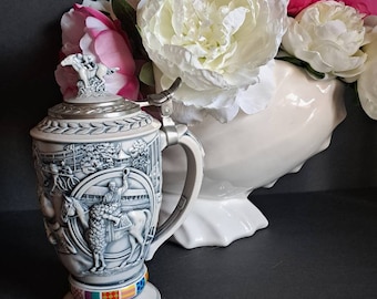 May include: A detailed, gray and white ceramic beer stein with a hinged lid and handle. The stein features a relief design depicting a horse race scene. The base has a colorful band. White and pink flowers are in the background.