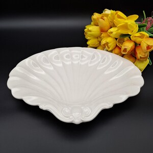 May include: A white, shell-shaped decorative dish with a scalloped edge and ornate detailing. The dish is made of ceramic and sits against a black background, with a bouquet of yellow tulips in the background.