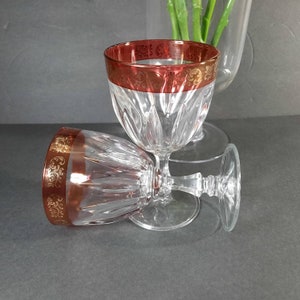 May include: Two clear glass wine glasses with a red and gold rim. The glasses are stacked on top of each other.