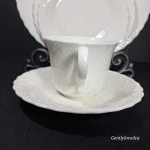K&ouml;nnte beinhalten: Wei&szlig;es Porzellan-Teetassen- und Untertassen-Set mit einem zarten Blumenmuster. Die Tasse hat eine abgerundete Form und einen einzelnen Henkel. Die Untertasse hat einen gewellten Rand.