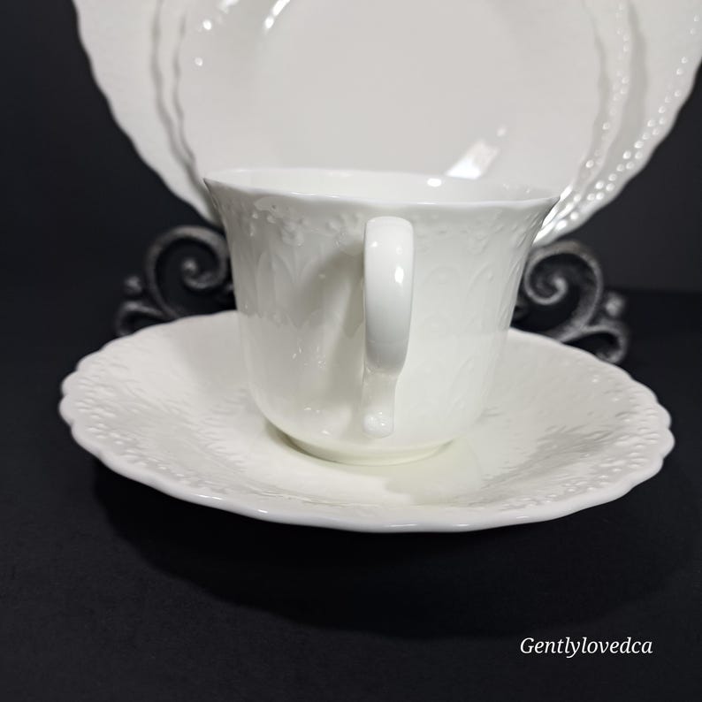 K&ouml;nnte beinhalten: Wei&szlig;es Porzellan-Teetassen- und Untertassen-Set mit einem zarten Blumenmuster. Die Tasse hat eine abgerundete Form und einen einzelnen Henkel. Die Untertasse hat einen gewellten Rand.