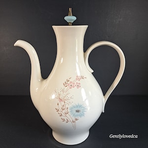 May include: A white ceramic teapot with a floral design in shades of pink, blue, and brown. The teapot has a lid with a blue knob and a handle on the side.