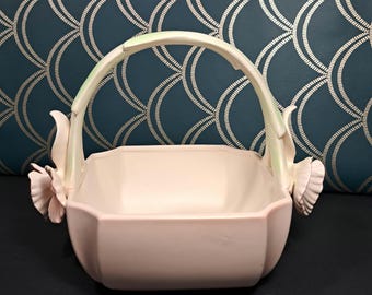 May include: A pale pink ceramic basket with a smooth matte finish, featuring a curved handle and sculpted floral accents on both sides. The basket has a square base with rounded corners and a light green tint on the top of the handle.