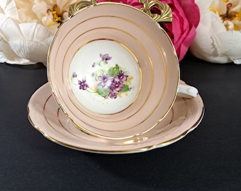 Peut inclure: Une tasse à thé et une soucoupe roses avec un motif floral délicat représentant des violettes violettes. La tasse et la soucoupe ont une bordure dorée.