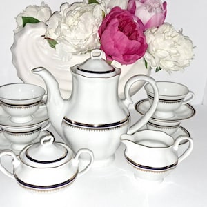 May include: A white porcelain tea set with gold and navy blue trim. The set includes a teapot, sugar bowl, creamer, and several teacups and saucers. A vase of white and pink peonies is in the background.
