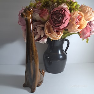 May include: A brown and black ceramic giraffe figurine with a long neck and a striped pattern. The figurine is standing on a white surface in front of a vase of pink and peach flowers.