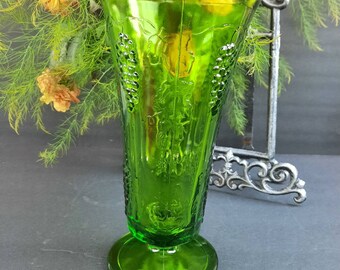 May include: A green glass vase with a textured grape design. The vase has a flared top and a round base.