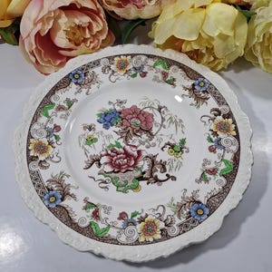 May include: A white ceramic dinner plate with a brown, green, yellow, pink, and blue floral pattern. The plate has a scalloped edge.