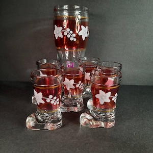 May include: Set of six clear glass shot glasses with red and white floral designs. The glasses have a boot-shaped base and are arranged in a group with a larger glass decanter in the background.