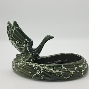 May include: A vintage green ceramic ashtray in the shape of a goose with outstretched wings. The ashtray has a dark green base with white crackle detailing. The goose's head and wings are also dark green.