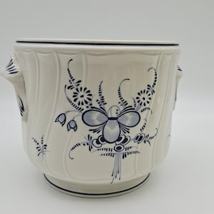 May include: A white ceramic planter with blue floral patterns. The planter has a rounded shape with a flared rim and two small handles. The blue floral design includes flowers, leaves, and other decorative elements.