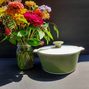 May include: A green ceramic casserole dish with a white lid and a knob on top. The dish is sitting on a table with a bouquet of flowers in a glass jar.