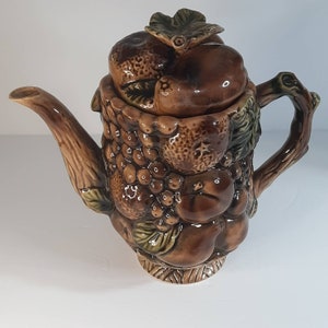 May include: A brown ceramic teapot with a lid shaped like a fruit basket. The teapot is decorated with brown and green glazed fruit and leaves.