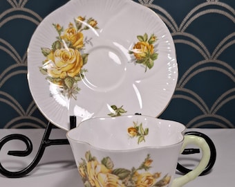 May include: A white teacup and saucer with a yellow rose pattern. The cup has a scalloped edge and a light green handle. The saucer has a scalloped edge and a gold trim.