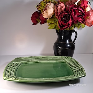 May include: A green ceramic serving platter with a bamboo design around the edges. The platter is rectangular with rounded corners. A black jug with a bouquet of red and pink flowers is in the background.