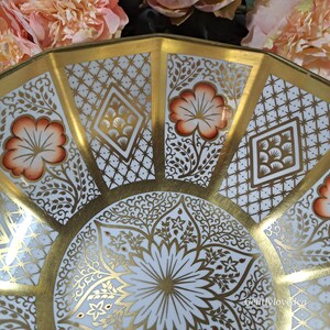 May include: A white porcelain bowl with a gold rim and intricate gold floral and geometric designs. The bowl has a scalloped edge and a white background.