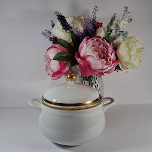 May include: White porcelain soup tureen with a gold trim and a gold knob on the lid. The tureen is sitting on a white surface in front of a bouquet of pink and white flowers.
