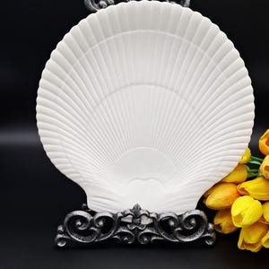 May include: A white, shell-shaped decorative dish with a ribbed design, displayed on a decorative, ornate, dark grey metal stand. A bouquet of yellow tulips is in the background.