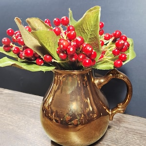 May include: A gold-colored pitcher with a handle, filled with a bouquet of red berries and green leaves. The pitcher has a shiny, reflective surface. The arrangement is set against a dark background.