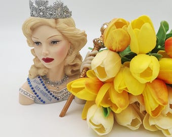 May include: A decorative bust of a woman with blonde hair, a silver crown, and a sash that reads "Miss All-America." A bouquet of yellow tulips is next to the bust. The bust is holding a scepter.