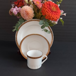 May include: A white teacup and saucer set with a gold rim. The saucer is nested inside a larger saucer, also with a gold rim. The set is on a black surface.